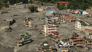 Aerial view of a town heavily impacted by flooding or a mudslide, with thick grey sediment covering streets and surrounding partially damaged buildings across a wide area.