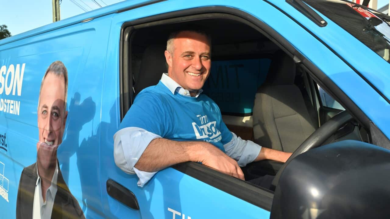 A man in a blue round-neck t-shirt worn over a shirt is sitting in the driver's seat of a blue vehicle, with one arm resting on the window.
