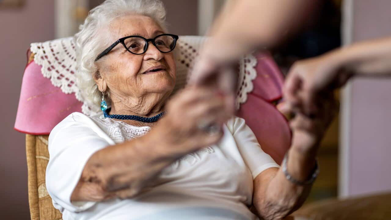 Female home carer supporting old woman to stand up from the armchair at care home