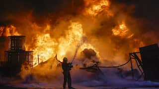 A silhouette of a firefighter spraying a high-pressure stream of water toward a massive, towering inferno that engulfs an industrial area at night.