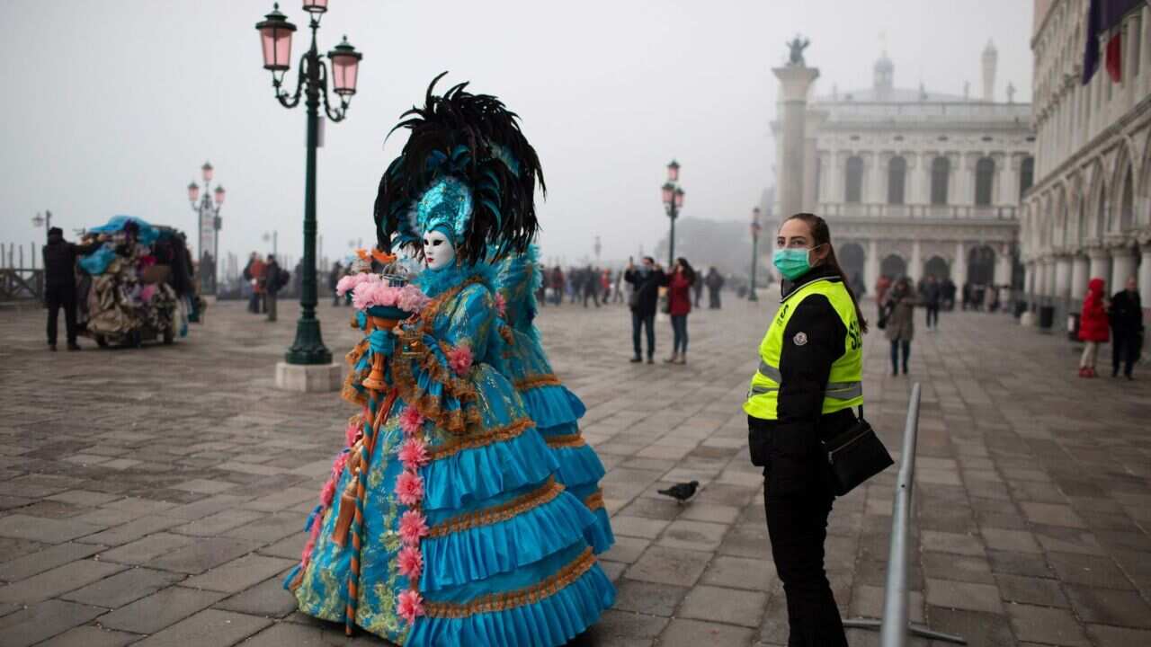 A security guard wearing a protective face mask stand guard as masked and costumed people walk by on the streets of Venice near San Marco square during the Carnival in Venice, Italy, 23 February 2020.