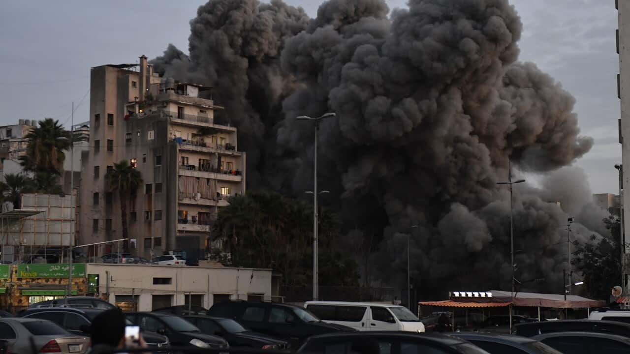 An apartment building in a city stands in the foreground, while a massive, dense plume of dark grey smoke billows into the sky directly behind it. The smoke is voluminous, suggesting a recent large-scale explosion or missile strike. In the lower foreground, a parking lot is filled with cars, and a few onlookers can be seen holding up their smartphones to record the scene. The sky is overcast and dim, adding to the somber and chaotic atmosphere of the event.