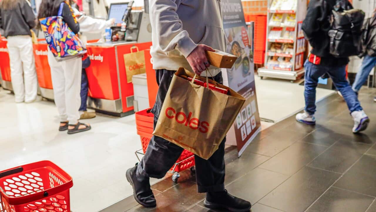 Torso view of a man carrying a brown paper Coles bag from a Coles supermarket. There are people using self-serve checkouts behind him