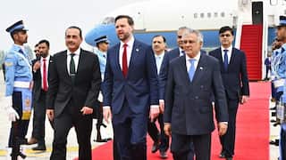 A group of government officials, including JD Vance and Ishaq Dar, walk along a red carpet on an airfield tarmac after disembarking from a US government aircraft, flanked by an honorary guard in blue uniforms.