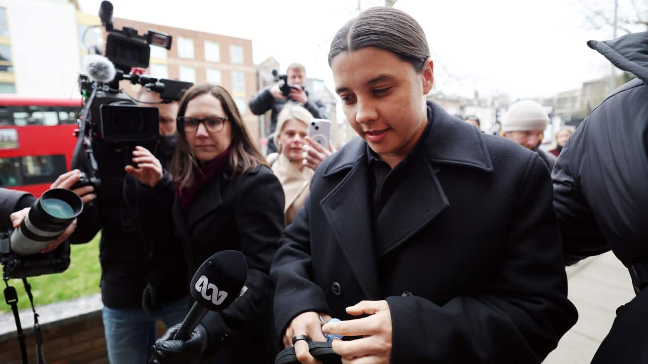 A woman in a black overcoat arrives at a trial mobbed by media.