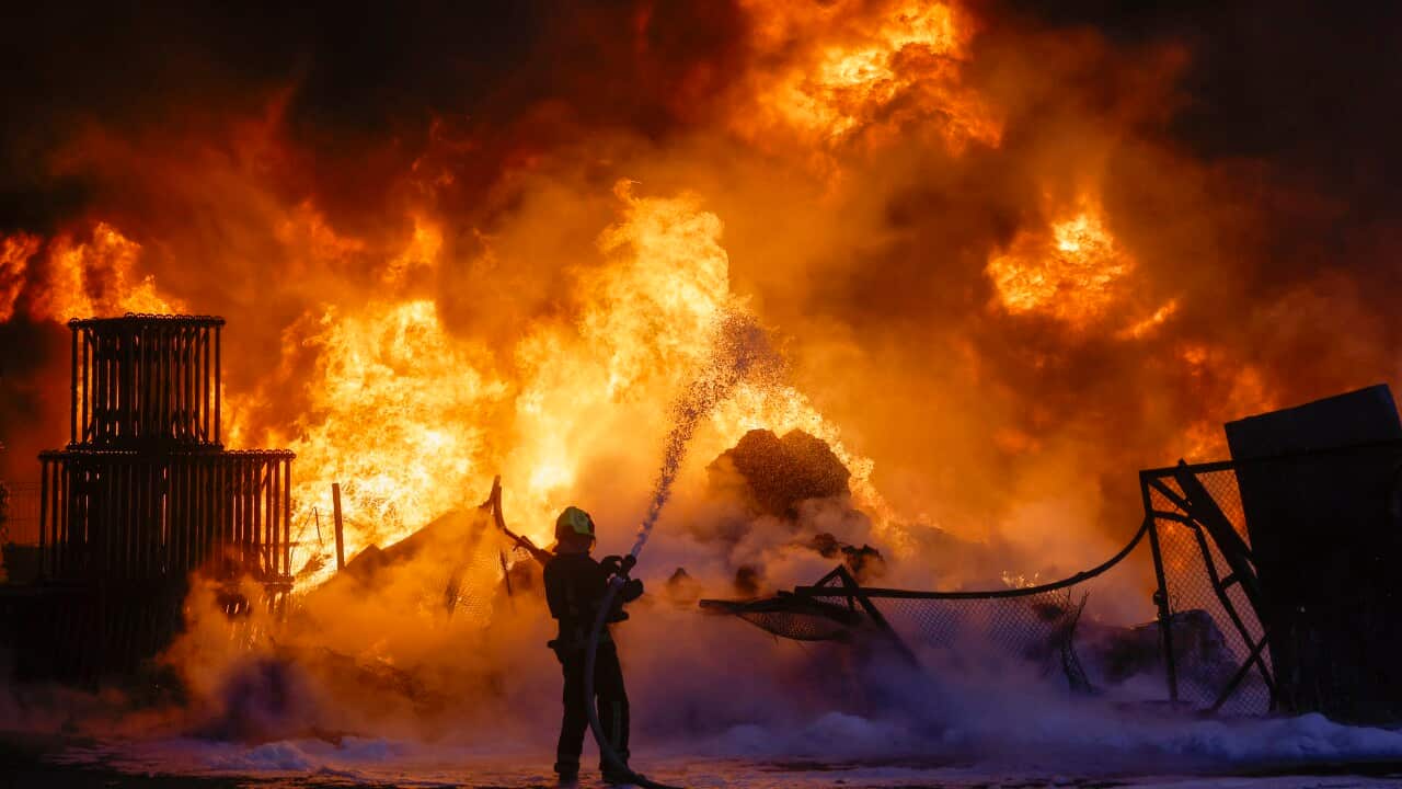 A silhouette of a firefighter spraying a high-pressure stream of water toward a massive, towering inferno that engulfs an industrial area at night.