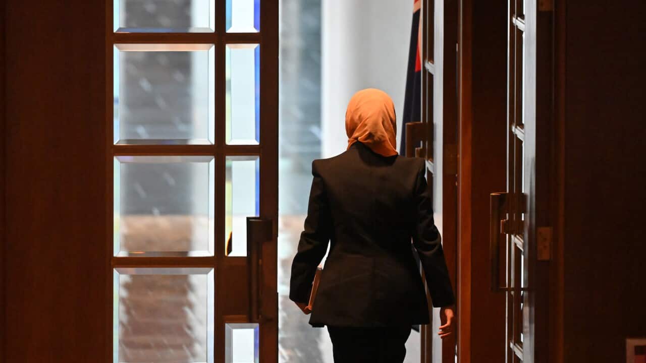 A woman (Fatima Payman) wearing a dark pantsuit and a light orange hijab walks out of the glass-panelled wooden doors of the Australian Senate
