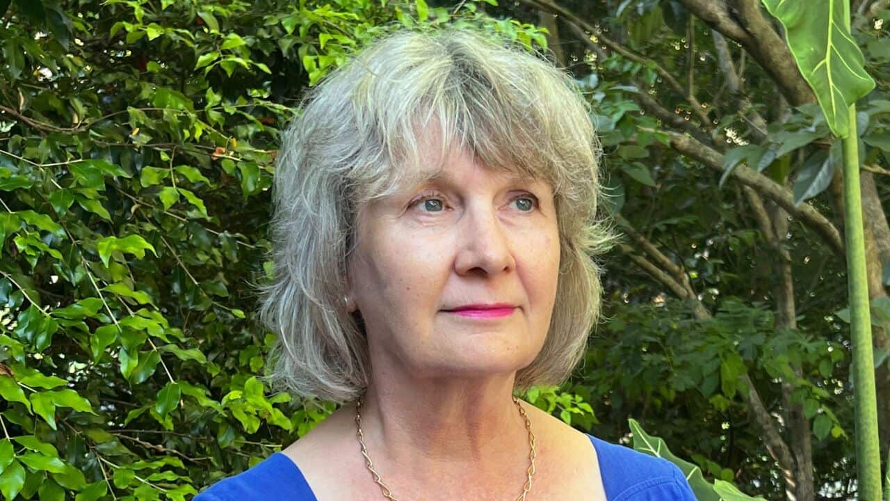 a close up of an older woman with ashy blonde grey hair in a blue shirt in front of tropical greenery