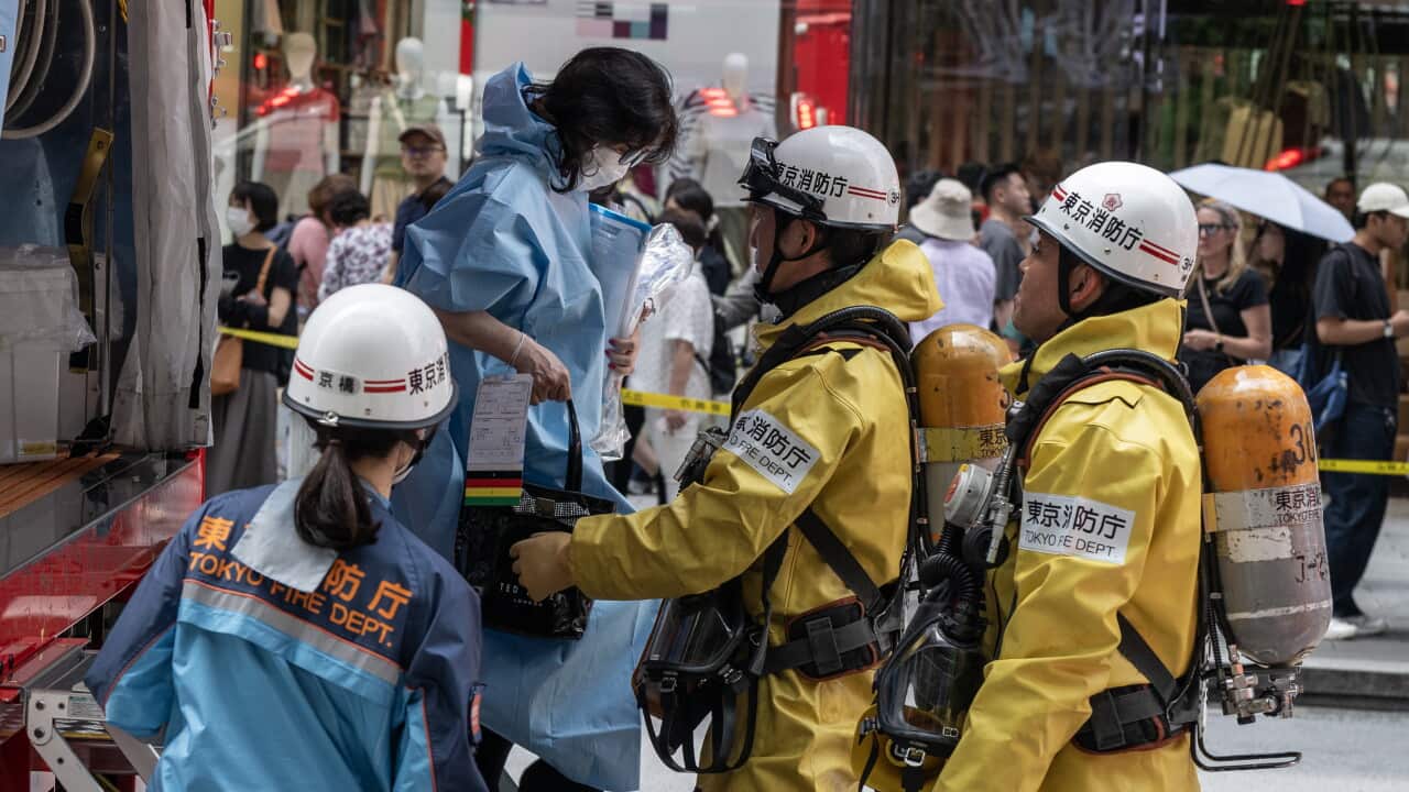A person wearing a yellow suit and a white helmet helps a woman wearing a blue suit to get down from an emergency vehicle. Another person in a yellow suit and white helmet and another in a blue suit and white helmet are watching. A crowd is seen in the background.