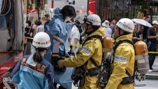 A person wearing a yellow suit and a white helmet helps a woman wearing a blue suit to get down from an emergency vehicle. Another person in a yellow suit and white helmet and another in a blue suit and white helmet are watching. A crowd is seen in the background.