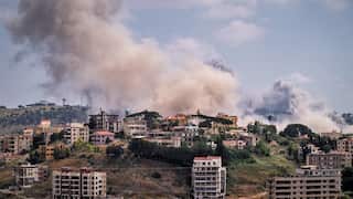 Smoke rises from suburban tower buildings amongst green hills and a blue sky.
