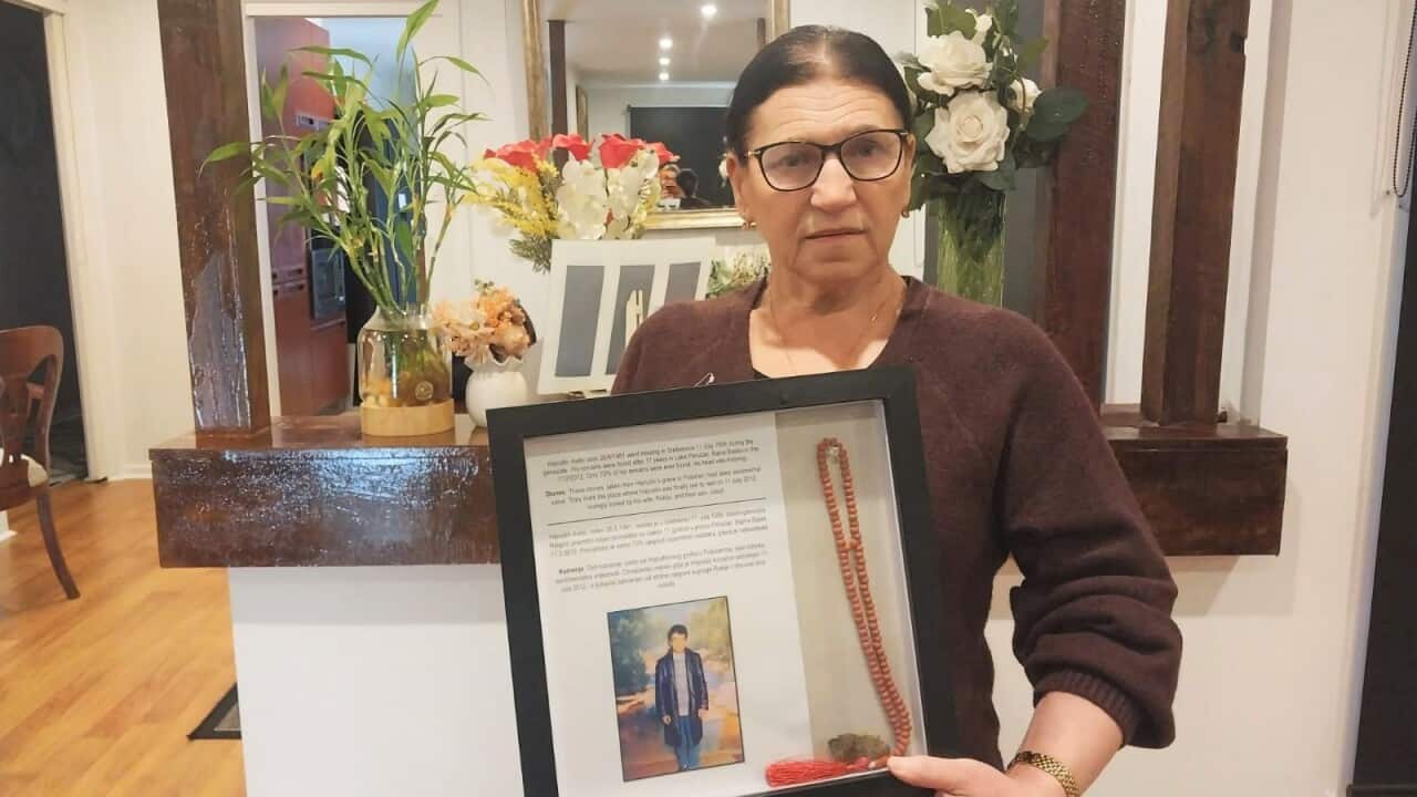 An older woman with dark hair and dark rimmed classes, wearing a brown jumper, holding up a frame containing the photo of a man, some writing, and a beaded necklace. Behind her is a mirror and flowers.