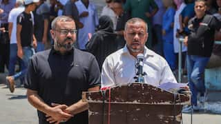 Two middle aged men stand behind a worn wooden podium. One is dressed in a dark blue shirt, the other in a white shirt. The man in the white shirt is speaking into a microphone on the podium.
