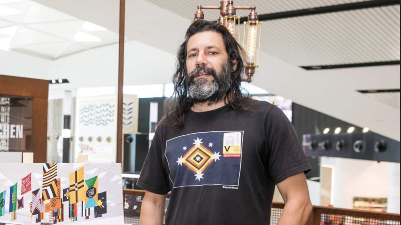 Artist Archie Moore stands in front of a photo of his United Neytions artwork in black jeans and t-shirt
