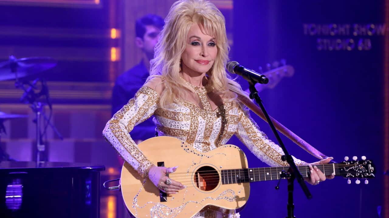 Person performing on stage with an acoustic guitar and microphone, wearing a white and gold embellished outfit under purple stage lighting.