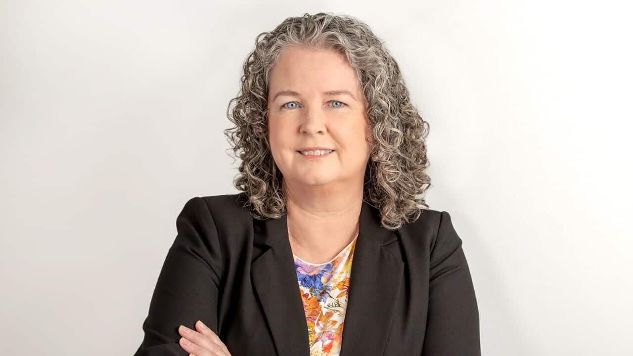 A middle-aged woman in a suit with curly grey hair smiles at the camera, her arms crossed.
