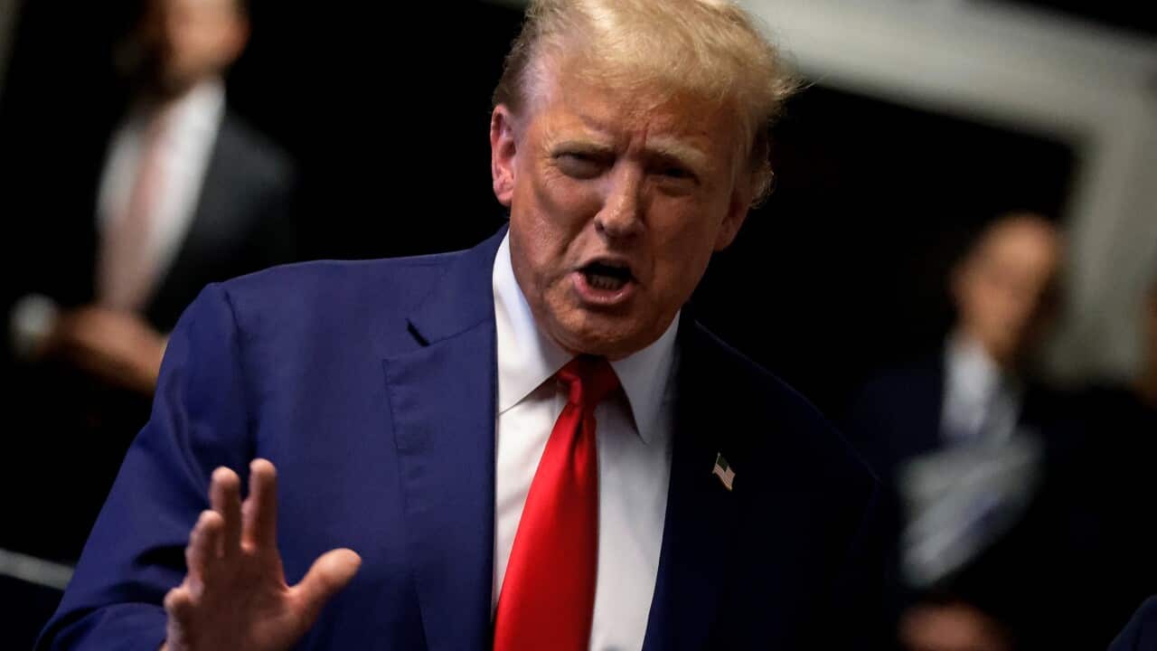 Donald Trump in a blue suit and red tie. He is speaking and holding his right hand at chest height.