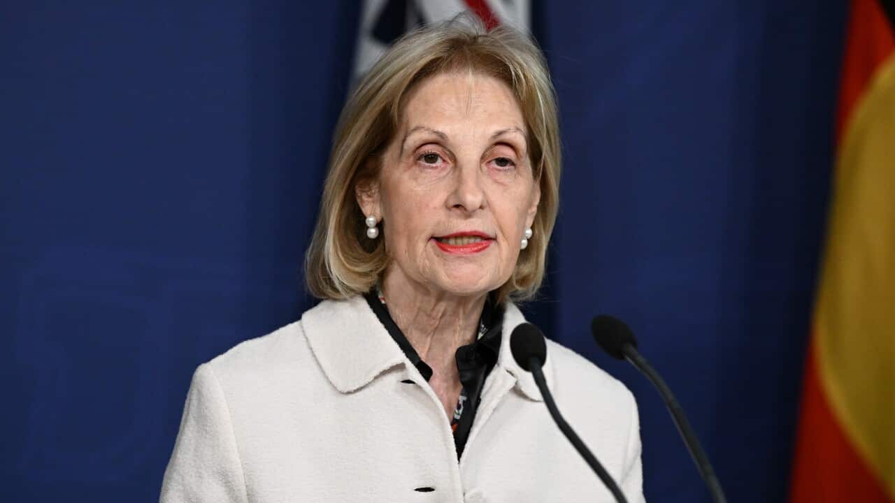 A woman with short blonde hair, wearing a white blazer, speaking into a microphone at a podium.