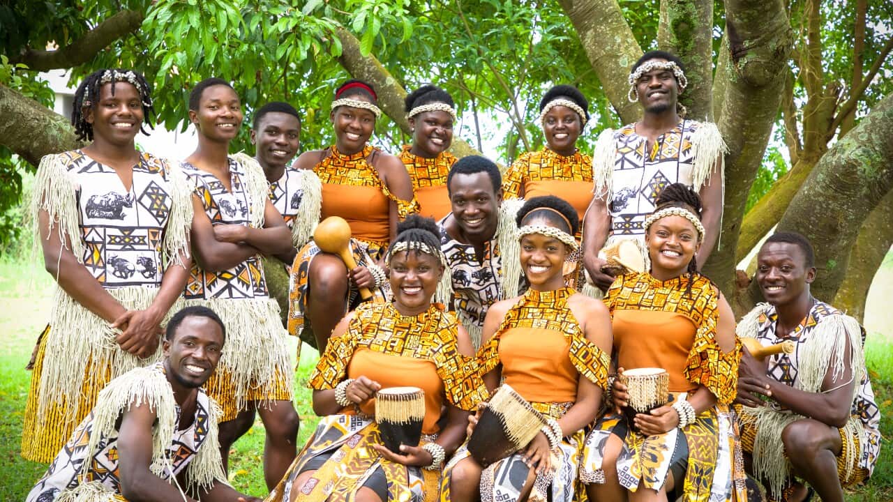 A group of African performers, some wearing white traditional costumes, and others orange traditional costumes, standing outside