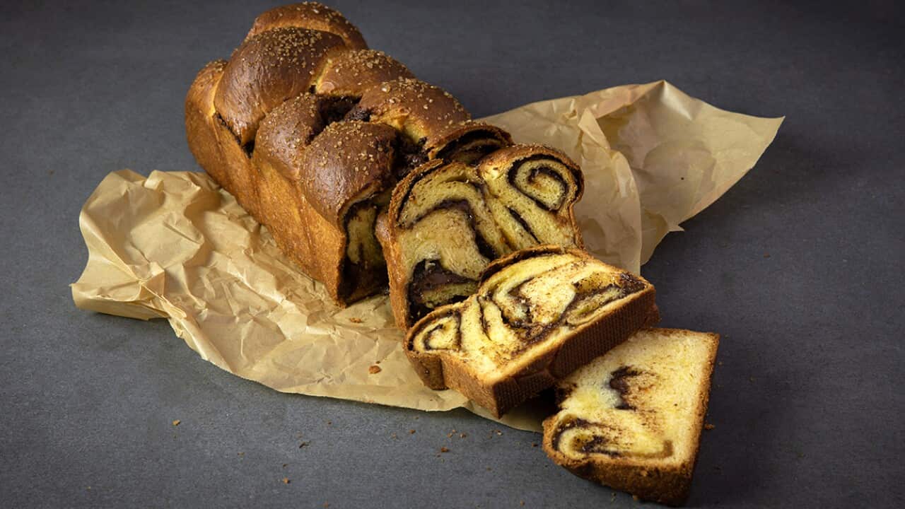 A loaf of golden plaited sweet bread sits on a piece of paper on a grey surface. The loaf has several slices cut from it, showing a swirl of chocolate running through the interior.