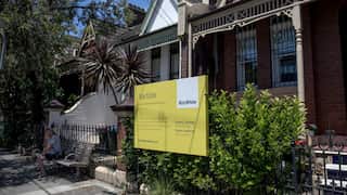 A Ray White auction sign stands outside a row of terrace houses in Sydney, with a person seated on a bench nearby.