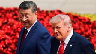 Two men in suits are lightly smiling as they walk in front of some red flowers.