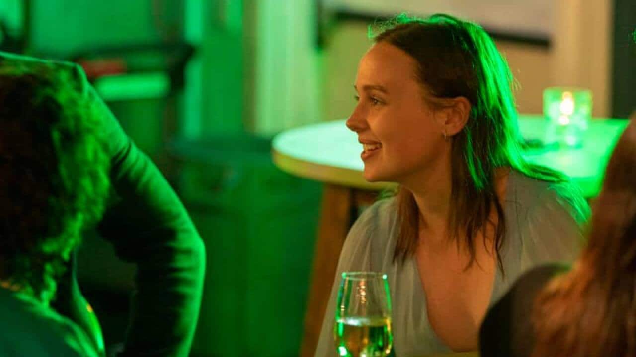 Woman with mid-length brown hair, waring a green dress. She's sitting at a table, not looking at the camera under green lighting. There is a wine glass on the table in front of her.