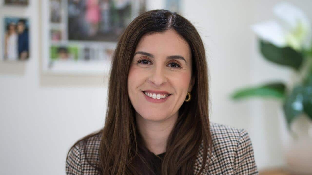 A woman with long brown hair and wearing a checkered blazer is smiling.