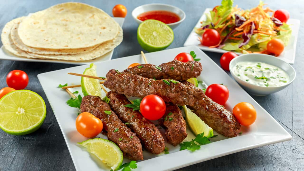 Homemade Kofta kebabs on skewers with pita, lime, vegetables, sweet chili and yogurt sauce on white plate.