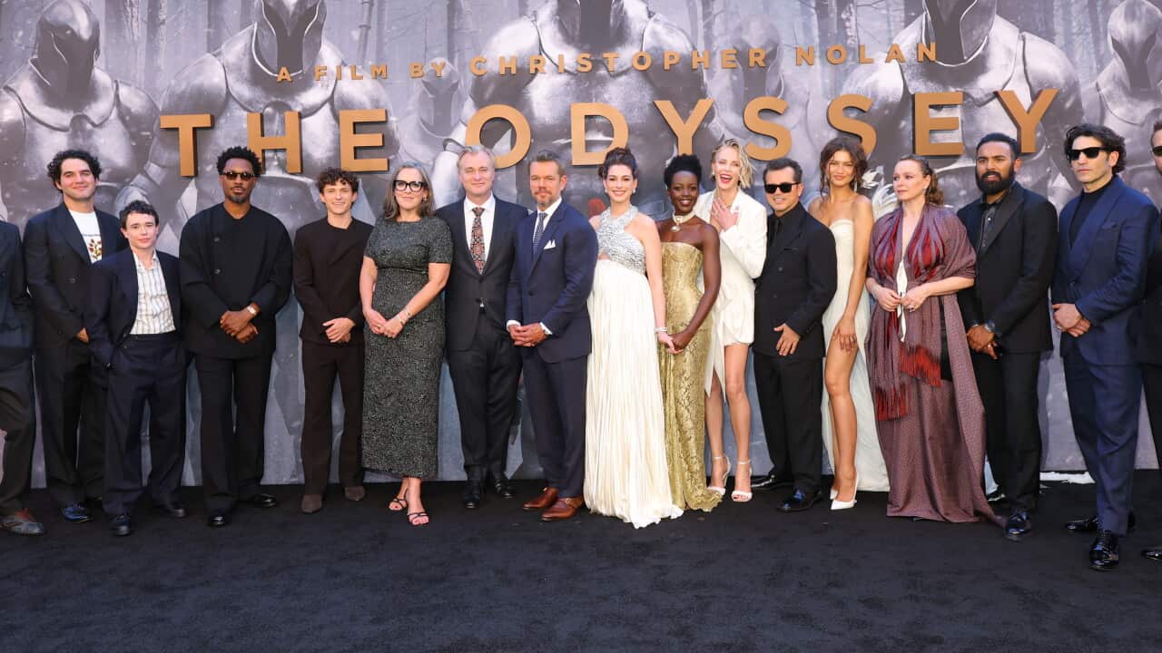 A photograph of the cast of the Odyssey at a premiere posing in front of a grey film poster on a blue carpet.