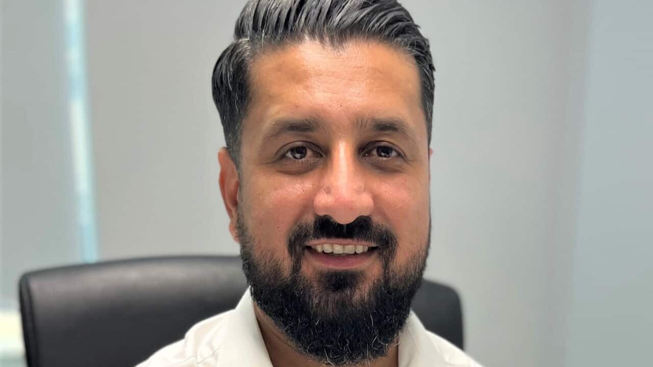 A man in a shite shirt sits at a desk smiling at camera.