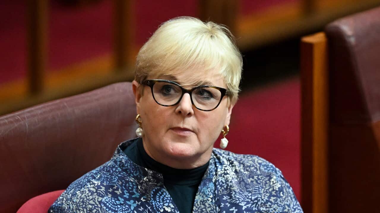 A woman with short blonde hair, glasses and heavy earings looks forward with a thoughtful expression on her face.