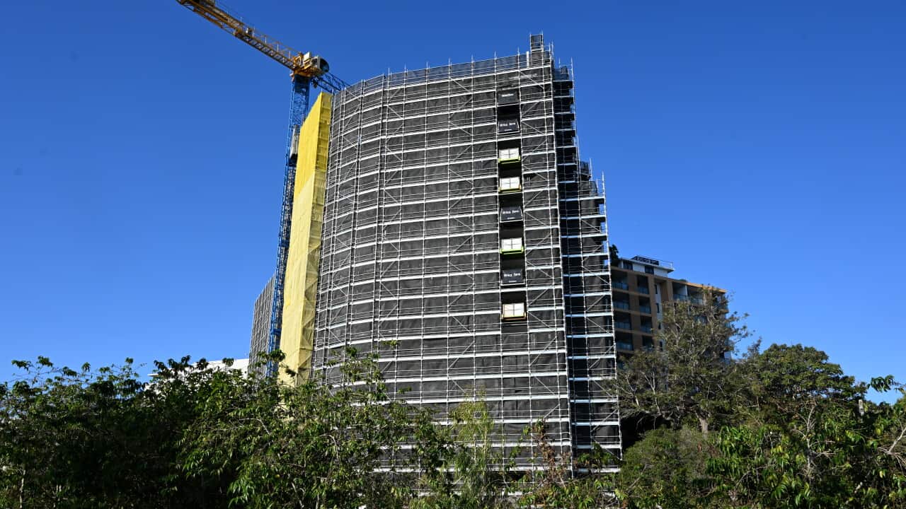 A tall building under construction on a clear day. A crane sits on the lefthand side of the building. Green trees sit at the bottom of the building.