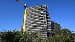 A tall building under construction on a clear day. A crane sits on the lefthand side of the building. Green trees sit at the bottom of the building.