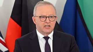 Australian Prime Minister Anthony Albanese speaks while wearing a dark suit, tie, and glasses, standing in front of Australian flags.