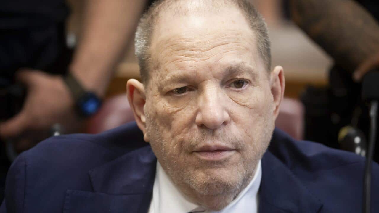 A man in a blue suit is sitting in a courtroom, with two guards standing behind him.
