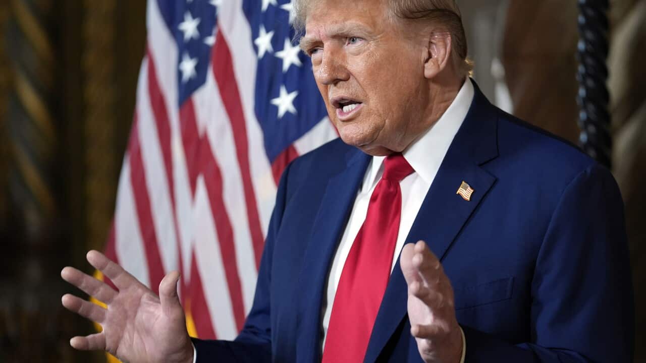 Sideways closeup of man in navy blue suit and red tie with hands up as he delivers a speech in front of an American flag.