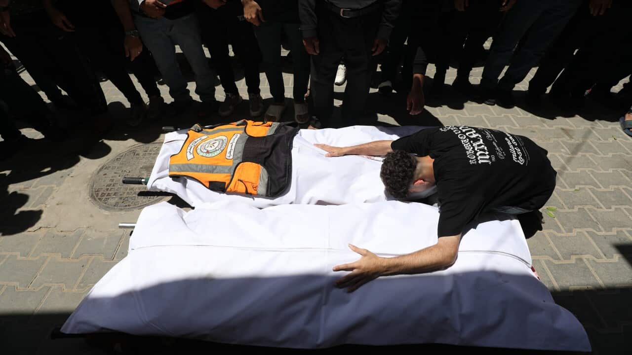 A man in a black t-shirt is grieving over the bodies of two people covered in white sheets. There's a crowd standing around him.