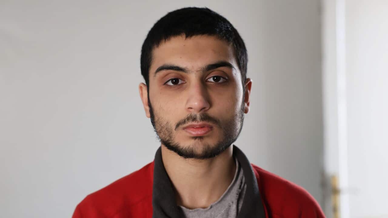 A young man with dark hair and a short beard wearing a grey t-shirt and a red zip-up jacket looks into the camera