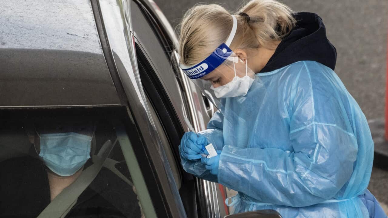 A woman wearing light blue personal protective top and a mask stands next to an open car window.
