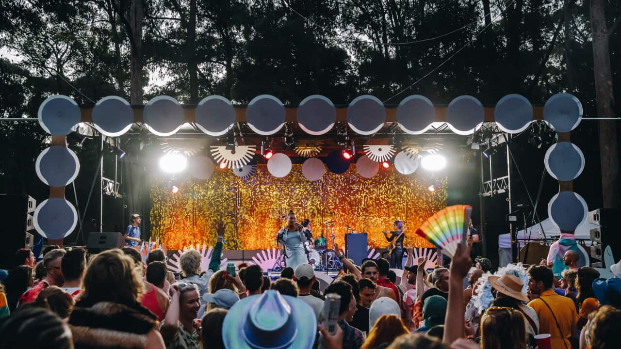 Seen from behind, a small crowd watches a stage on which glowing yellow confetti appears to be raining down upon a performer.
