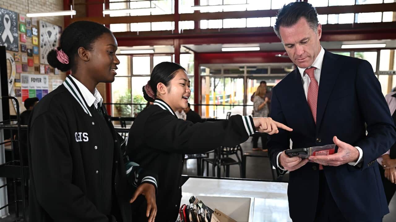 Two school students point to a phone carried by Chris Minns over a table with phones in pouches.