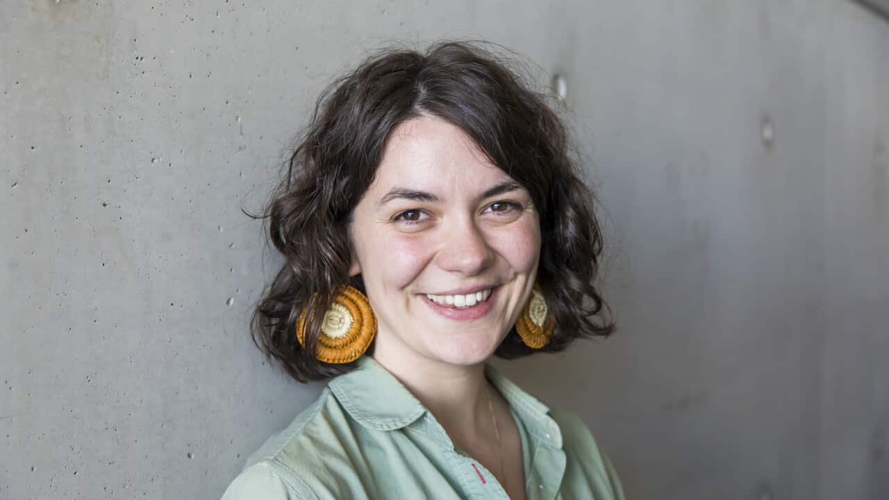 Jazz Money, smiling and standing in front of a grey wall wearing a green collared shirt and orange and yellow earrings.