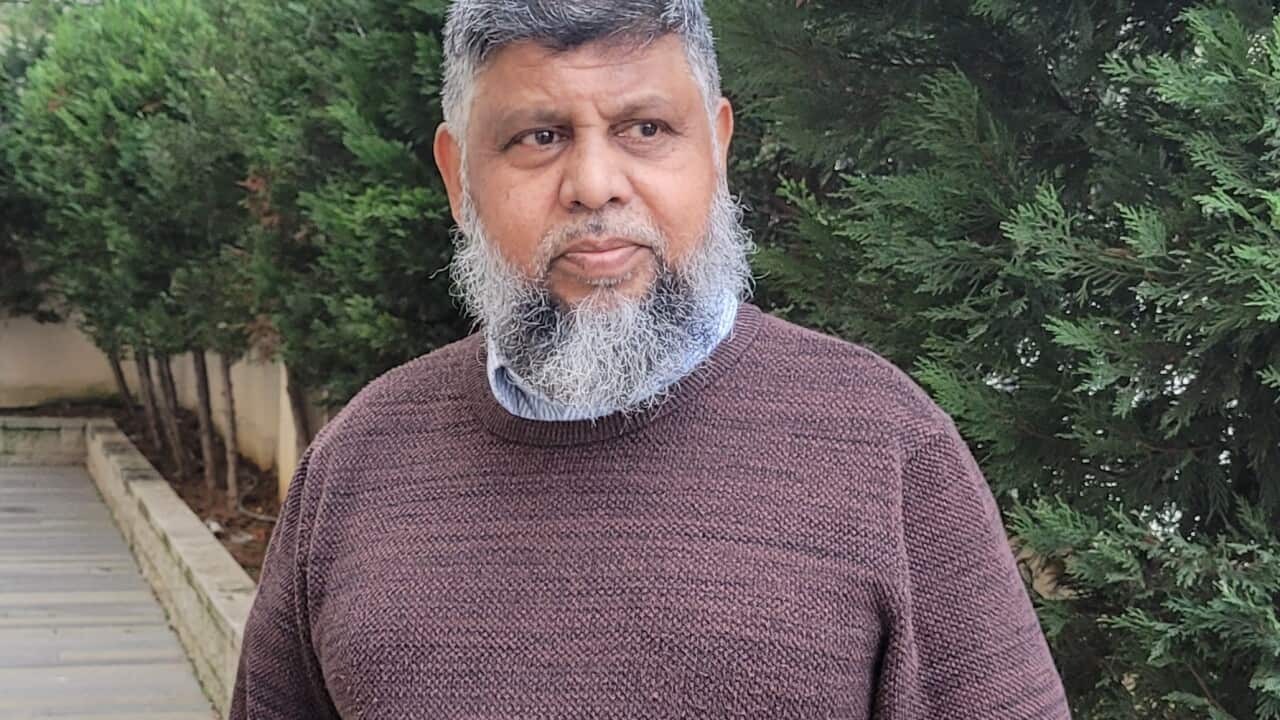 A man stands with hais hands in his pockets outside in a brown jumper.