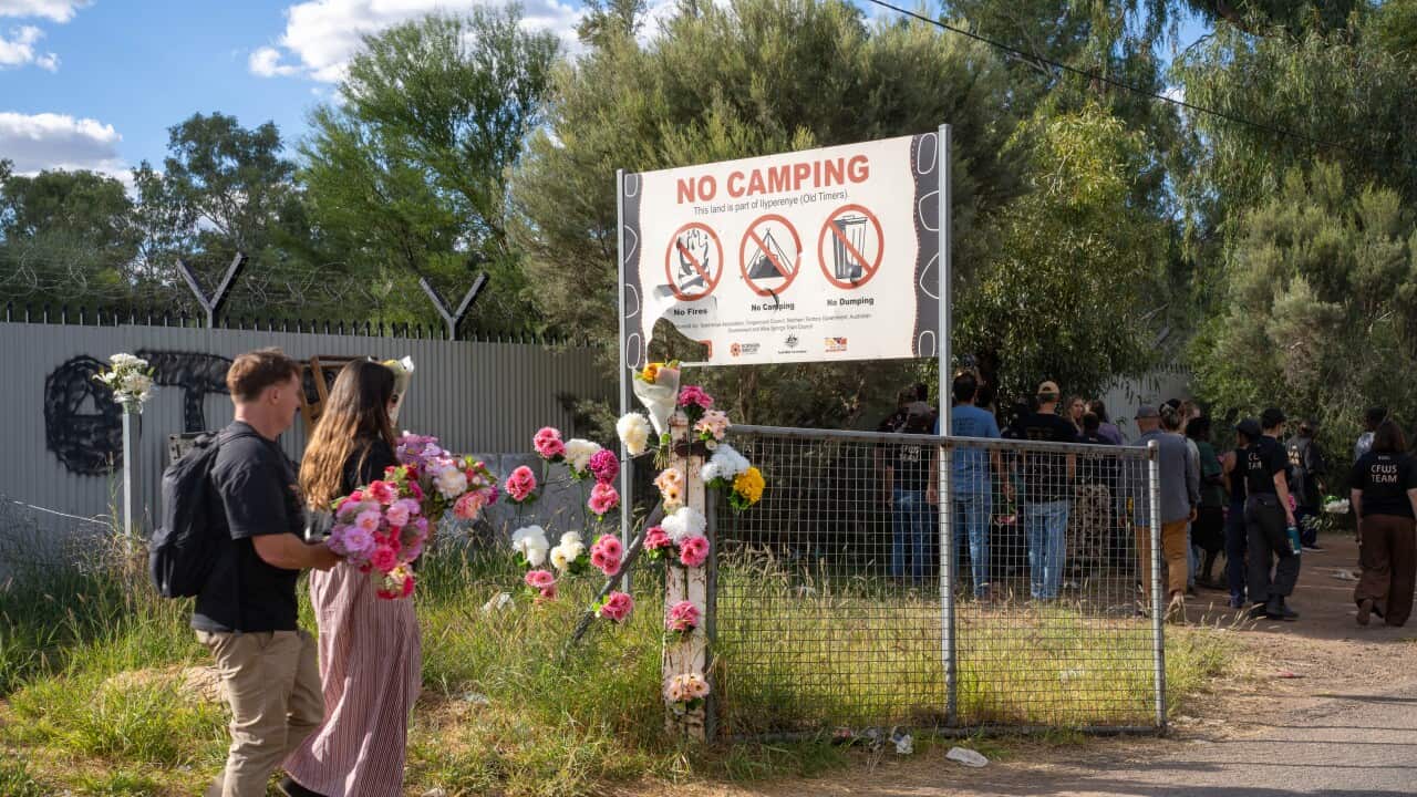 Two people walk with flowers in their hands towards a fence where flowers have been placed. There's a sign behind the fence that says, 'No Camping.'