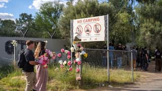 Two people walk with flowers in their hands towards a fence where flowers have been placed. There's a sign behind the fence that says, 'No Camping.'