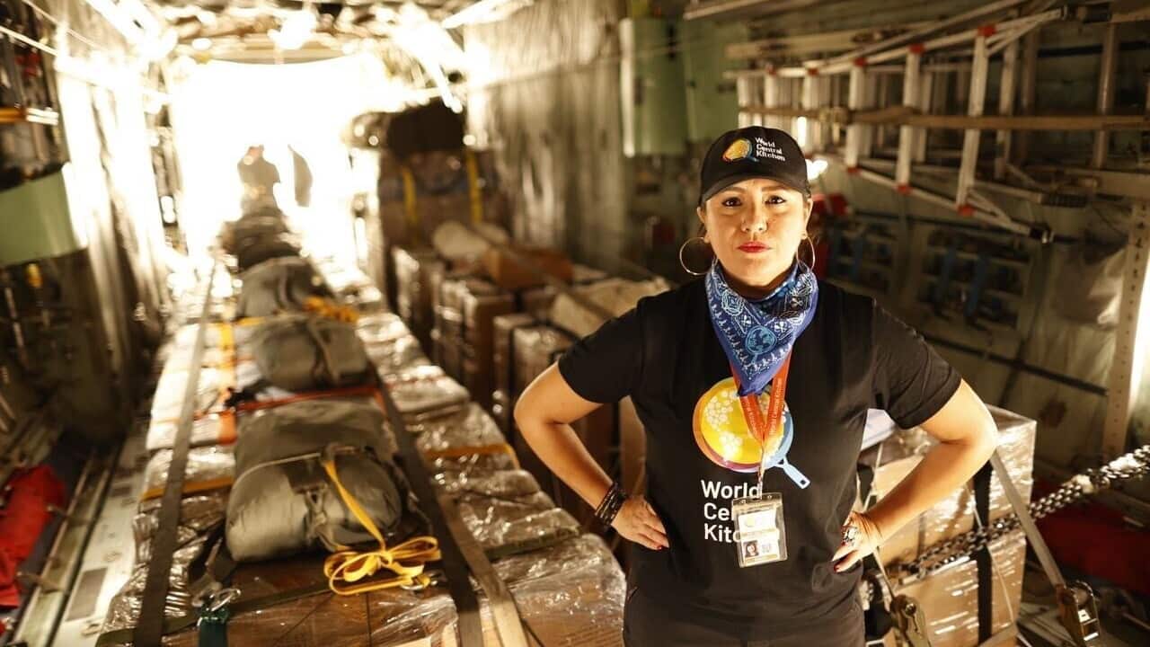 A woman with her arms on her hips, wearing a t-shirt and hat with the words 'World Certified Kitchen' on it. She's standing in front of aid packages.