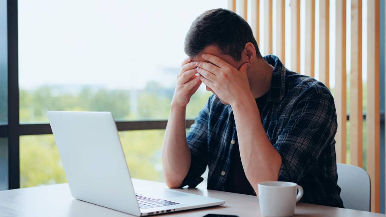 A man wearing a blue tartan shirt, sitting in front of a laptop with his head in his hands