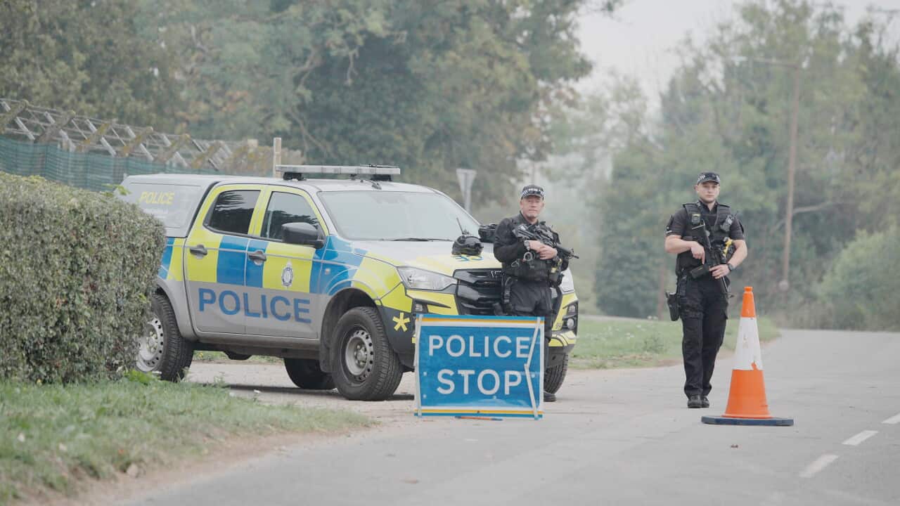 Police officers stand beside a marked police vehicle at a rural road checkpoint, with a “POLICE STOP” sign and traffic cone positioned on the roadway.