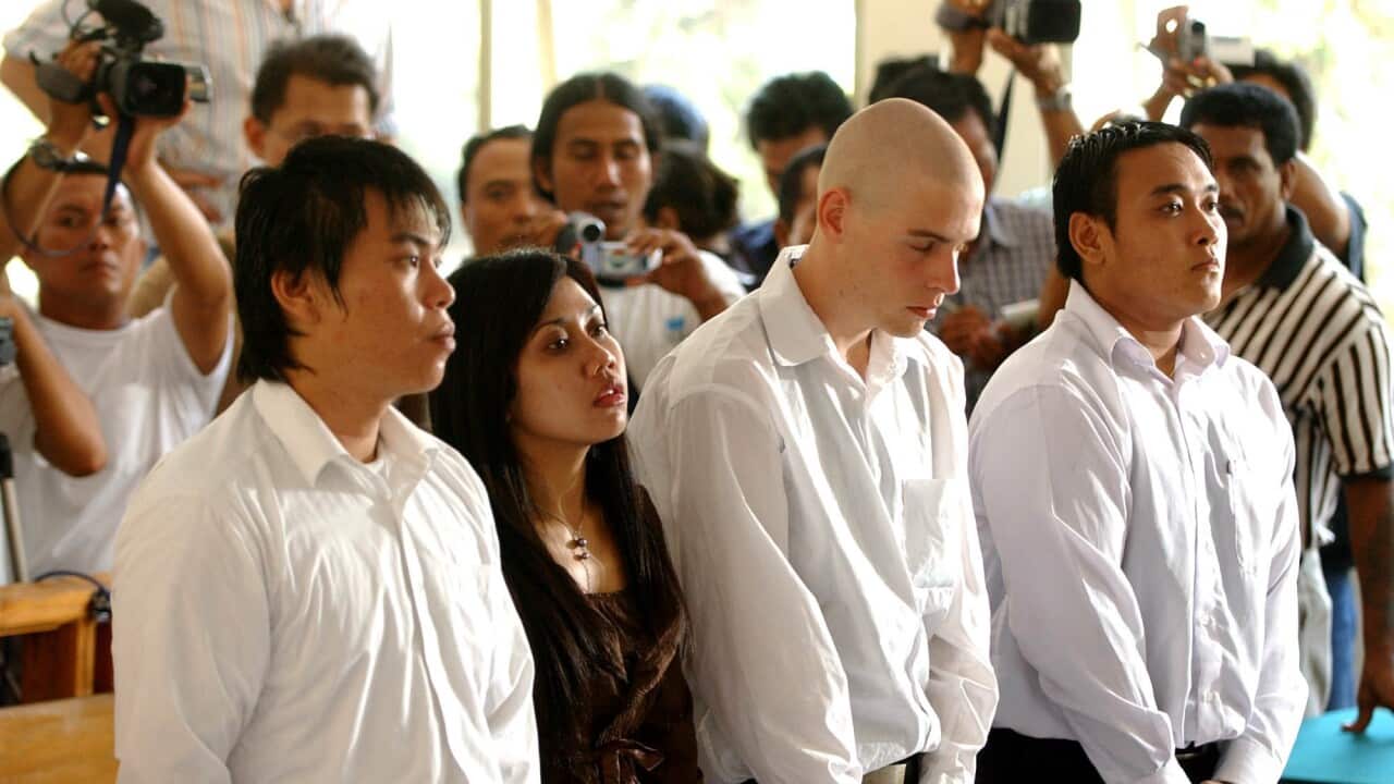 Three men in white shirts stand with a woman between them while media personnel stand behind them.